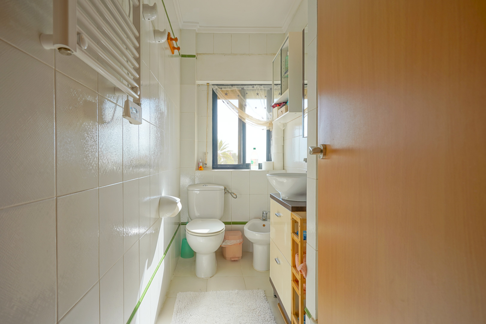 Functional bathroom featuring white tiling, a bidet, a toilet, and a vanity with a vessel sink. A window provides natural light and views of the outdoors.