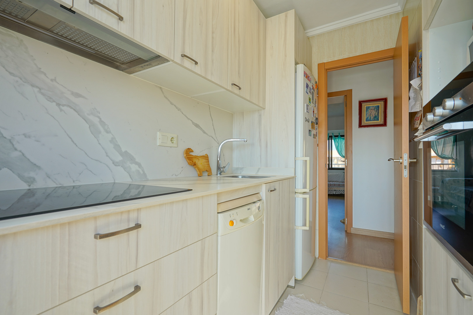 Functional kitchen featuring light wood cabinetry, a marble-effect backsplash, induction cooktop, and integrated appliances with a hallway view.