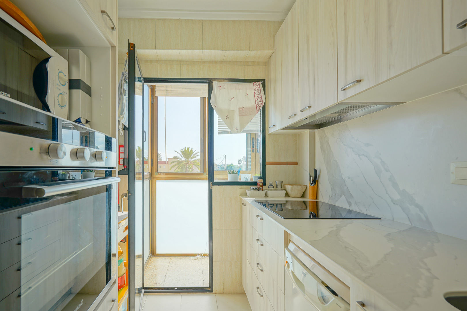 Functional kitchen featuring light wood cabinetry, marble-effect countertops, and a glass door leading to a balcony with palm tree views.