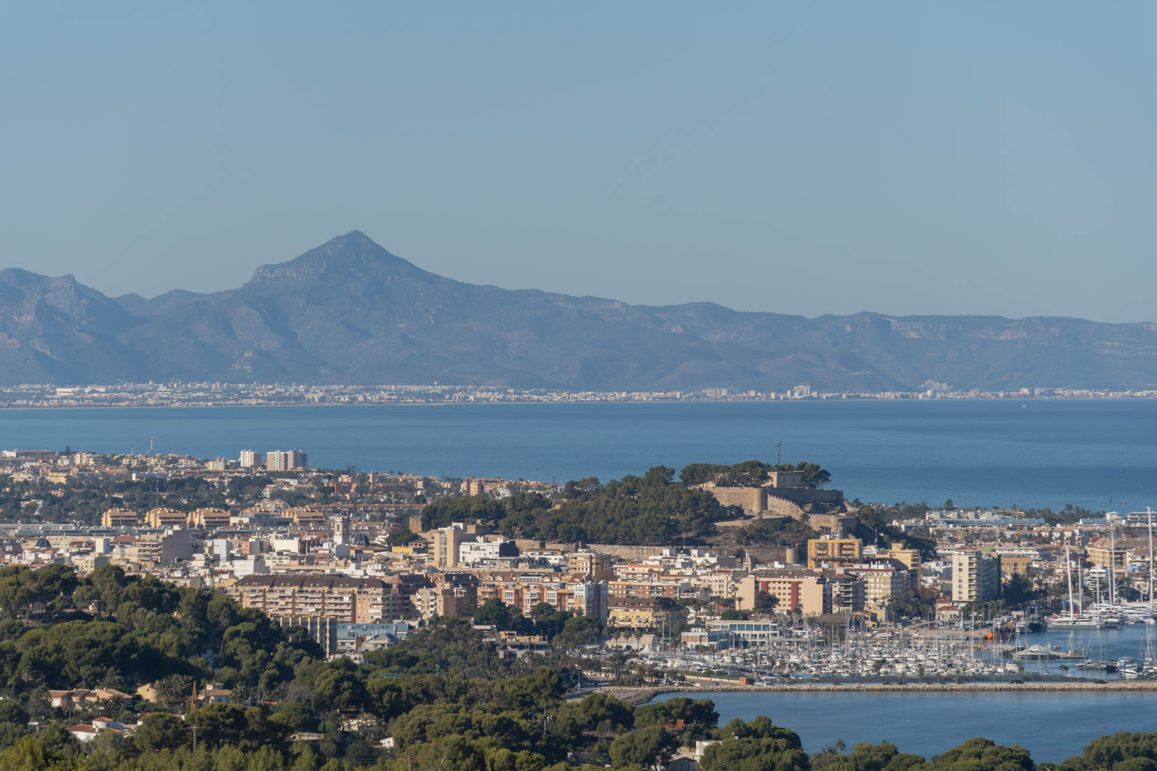 Panoramic view of the coastal city featuring the historic hilltop castle, the marina with numerous yachts, and a distant mountain range across the Mediterranean bay.