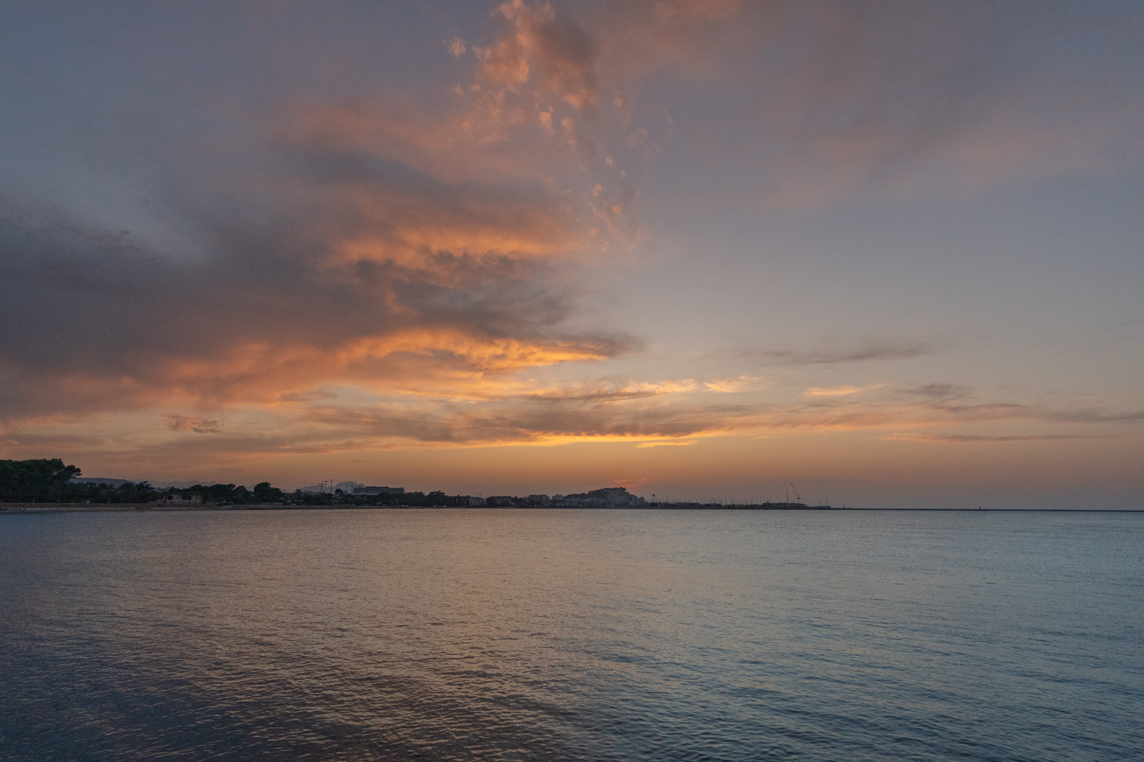 Panoramic view of the Mediterranean coastline at sunset, featuring orange-tinted clouds and calm waters reflecting the twilight sky over the distant town.