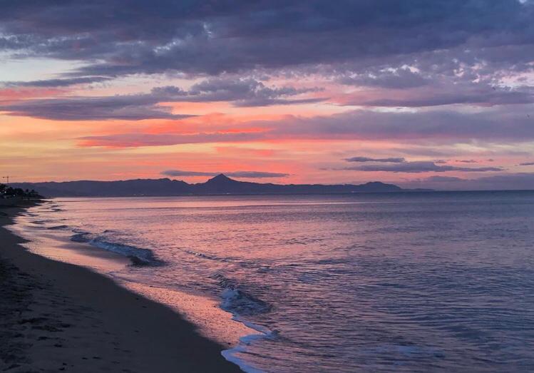 Panoramic view of the sandy coastline at dusk, featuring a mountain silhouette against a vibrant purple and orange sky.