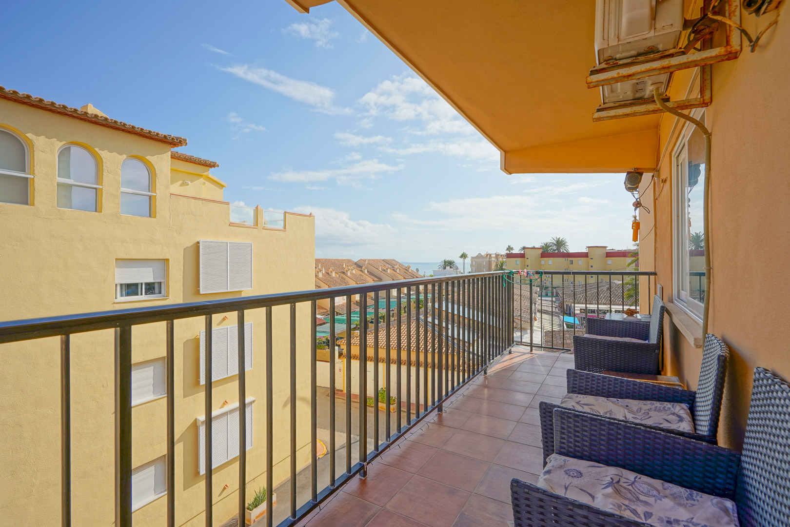 Private balcony featuring terracotta tile flooring, black metal railings, and wicker seating. It offers views of the surrounding architecture and a distant sea horizon.
