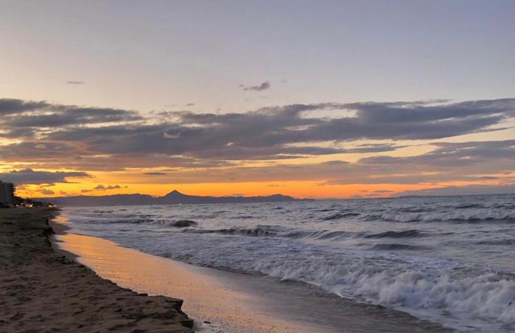 Sunset view over a sandy beach with crashing waves and distant mountain silhouettes under a cloudy orange sky.