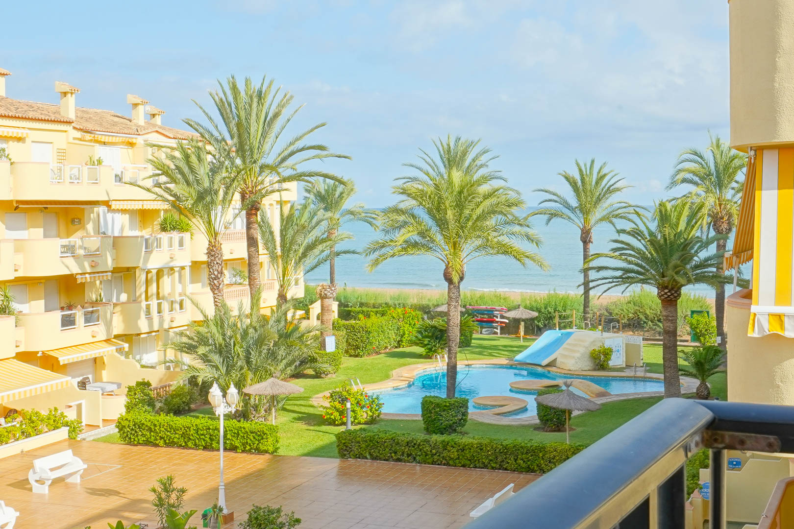 View from a private balcony overlooking a communal garden with palm trees, a swimming pool with a slide, and direct views of the Mediterranean Sea.