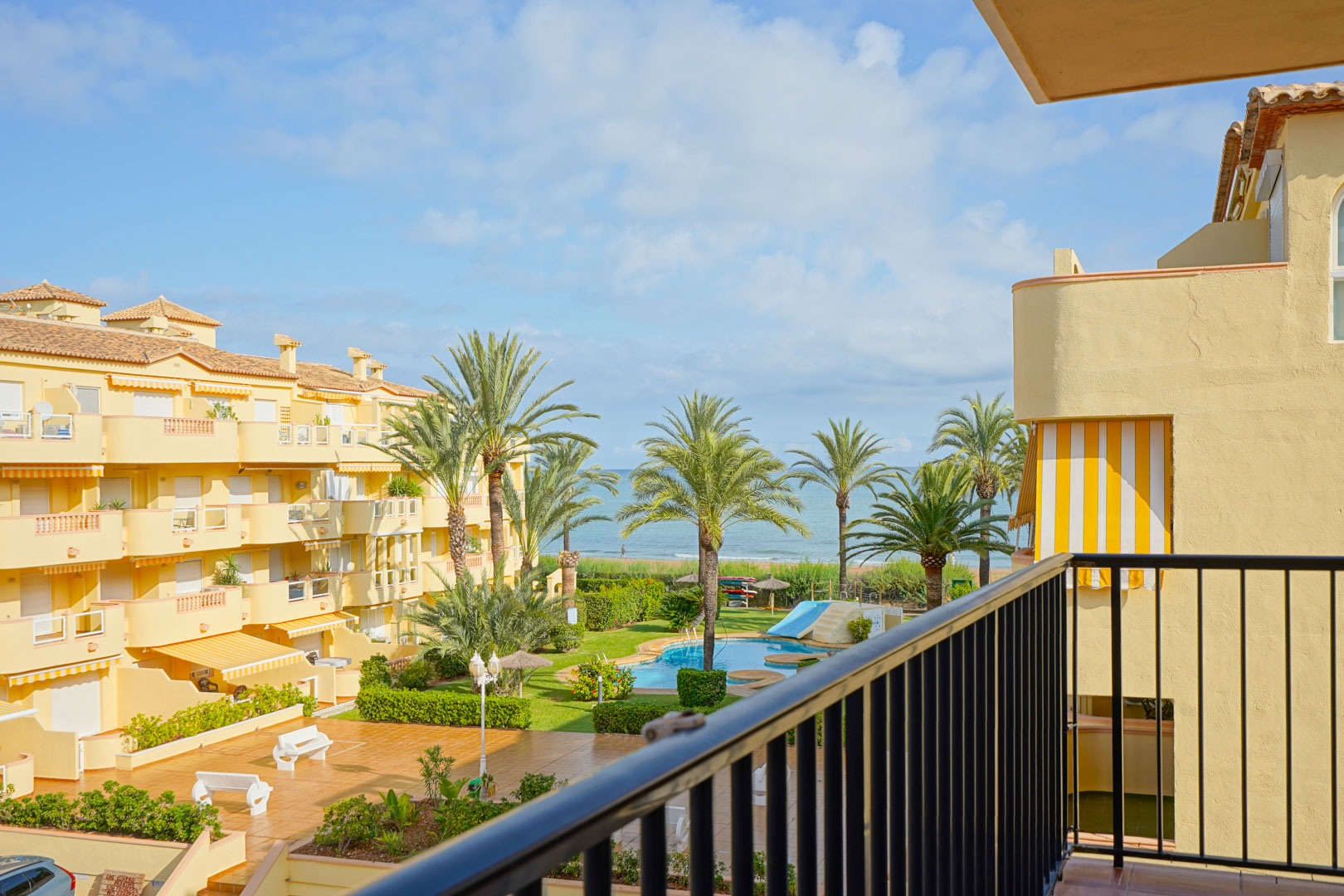 View from a private balcony overlooking a landscaped residential complex with a swimming pool, palm trees, and direct vistas of the Mediterranean Sea.