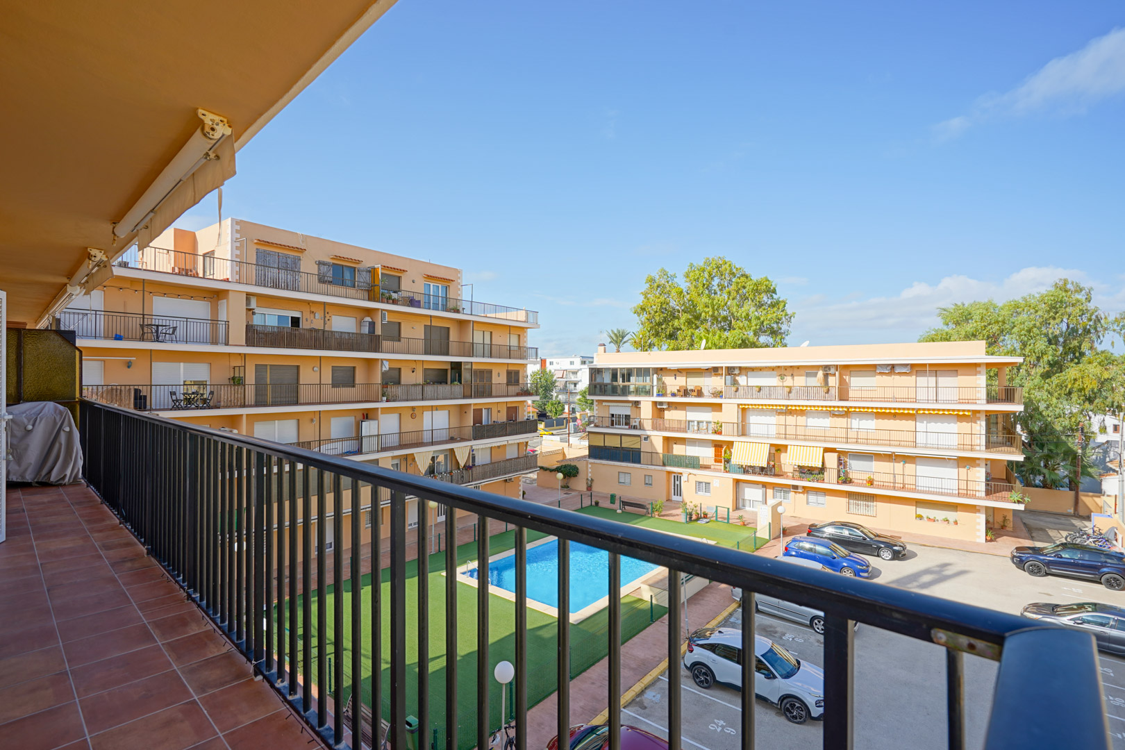 View from a private balcony with terracotta tiles and metal railing, overlooking a communal swimming pool, artificial lawn, and parking area within a residential complex.