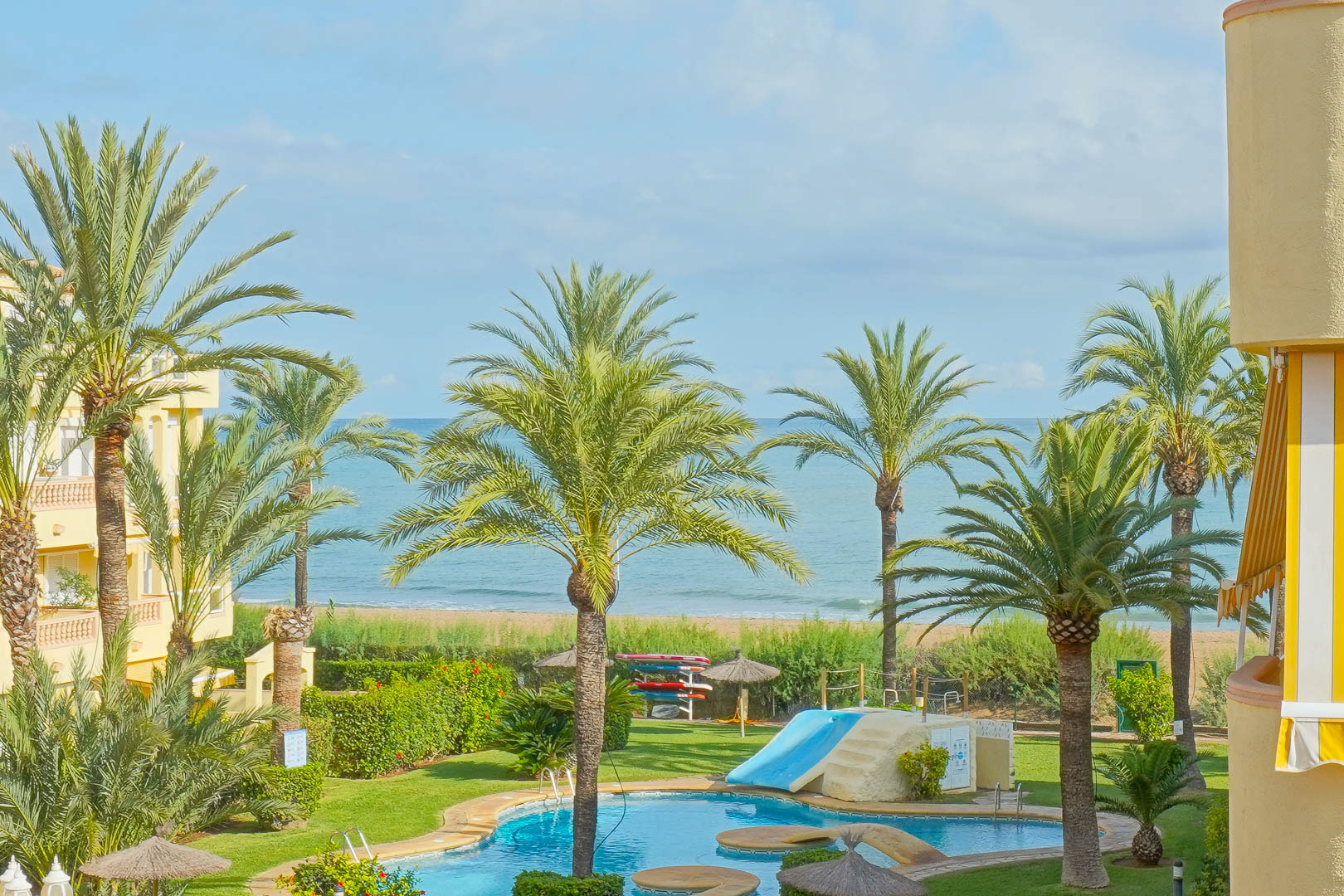 View of a communal swimming pool with a slide, surrounded by palm trees and green lawns, overlooking the sandy beach and the Mediterranean Sea.