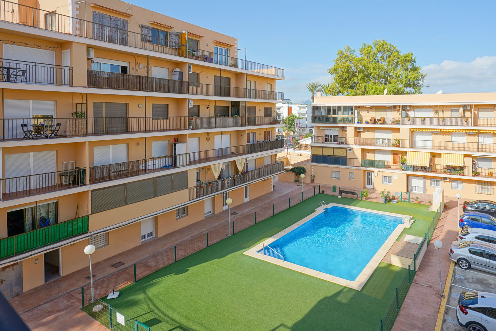 View of the residential complex featuring a rectangular swimming pool, fenced artificial turf area, and multi-story buildings with private balconies and parking.