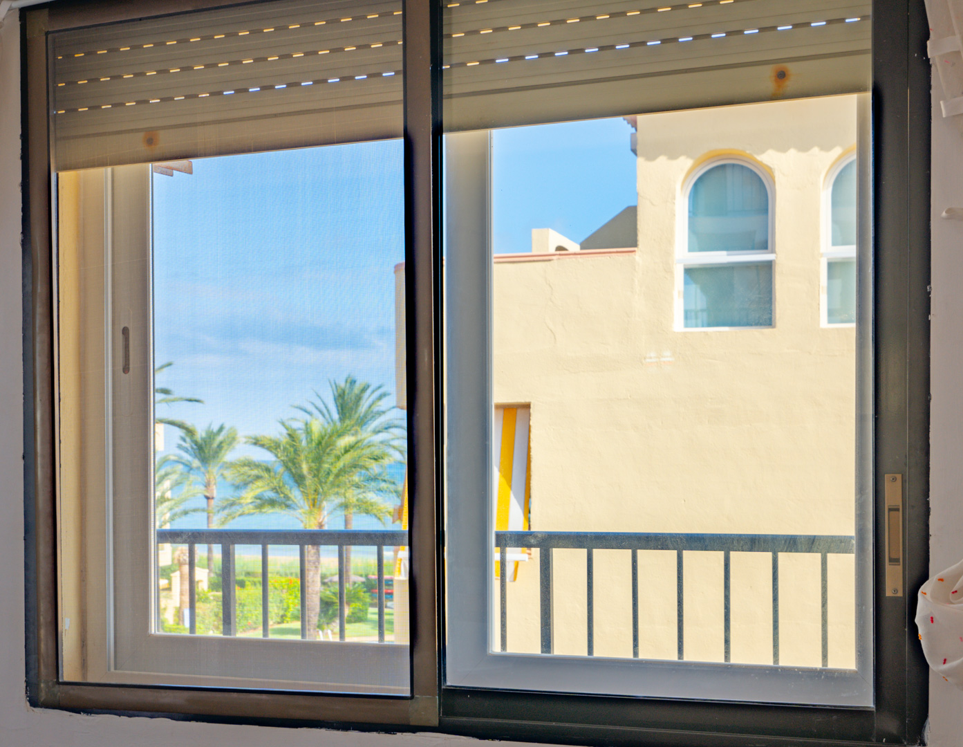 View through a sliding window with integrated roller blinds, showing palm trees, a glimpse of the sea, and Mediterranean architecture under a clear sky.