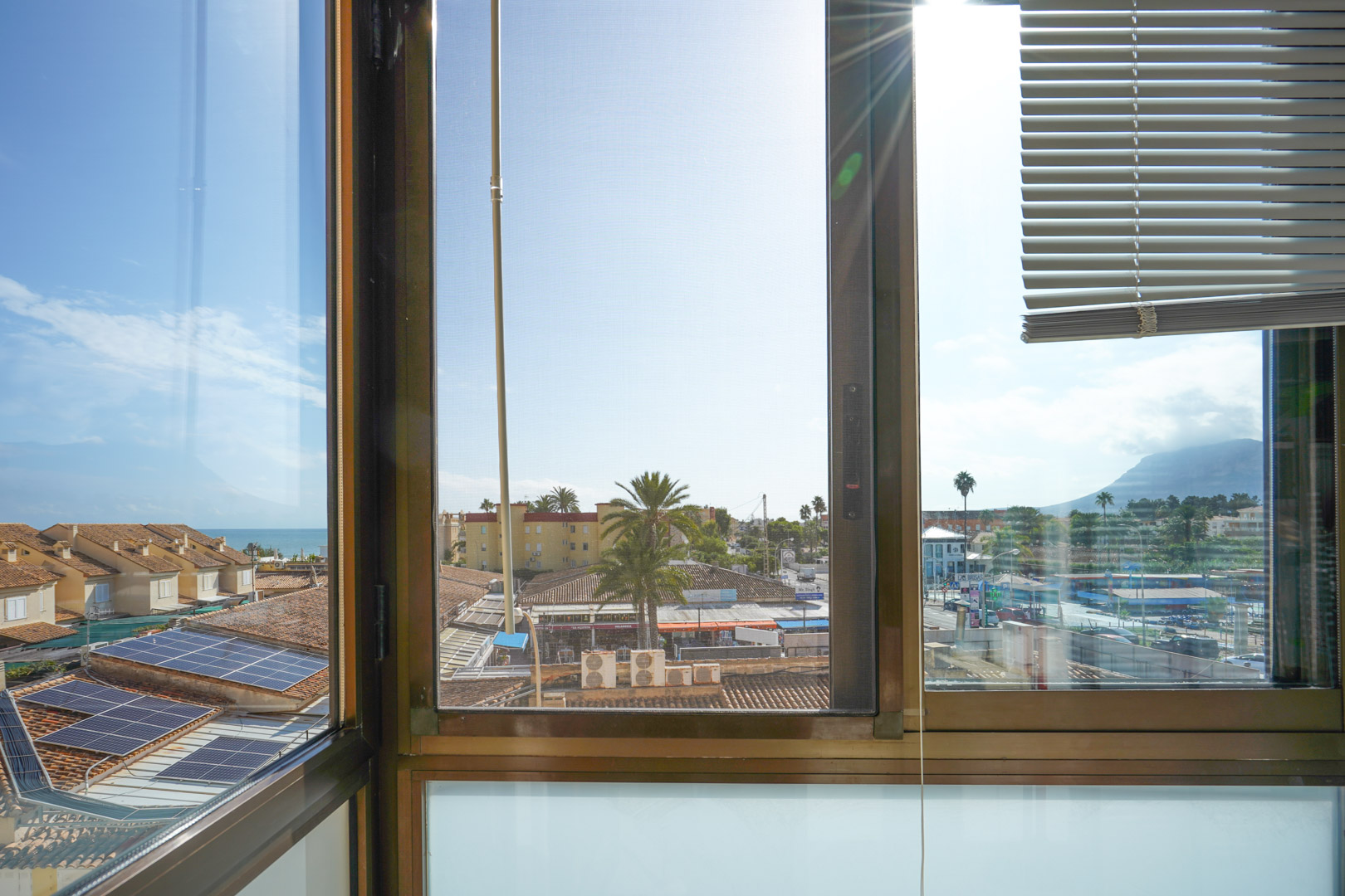 View through a window with blinds and fly screen, overlooking tiled roofs with solar panels, palm trees, and distant views of the sea and mountains.