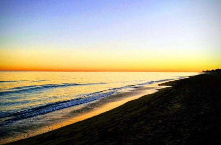 Wide-angle view of a sandy shoreline at dusk, featuring gentle waves and a gradient sky transitioning from deep blue to golden yellow over the horizon.
