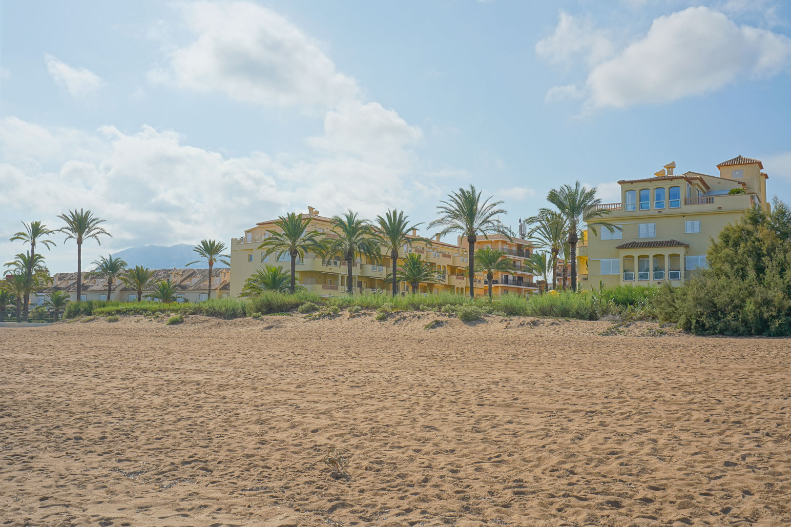 Wide sandy beach fronting Mediterranean-style residential buildings with palm trees and distant mountain views under a bright sky.
