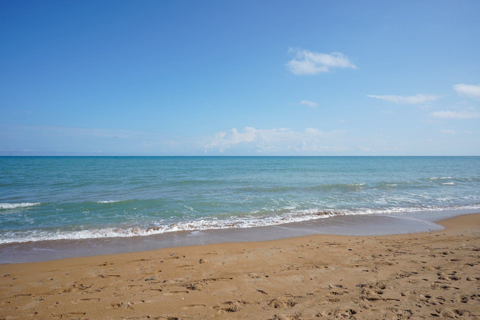 Wide sandy beach with gentle turquoise waves under a clear blue sky. The shoreline features golden sand and calm Mediterranean waters.