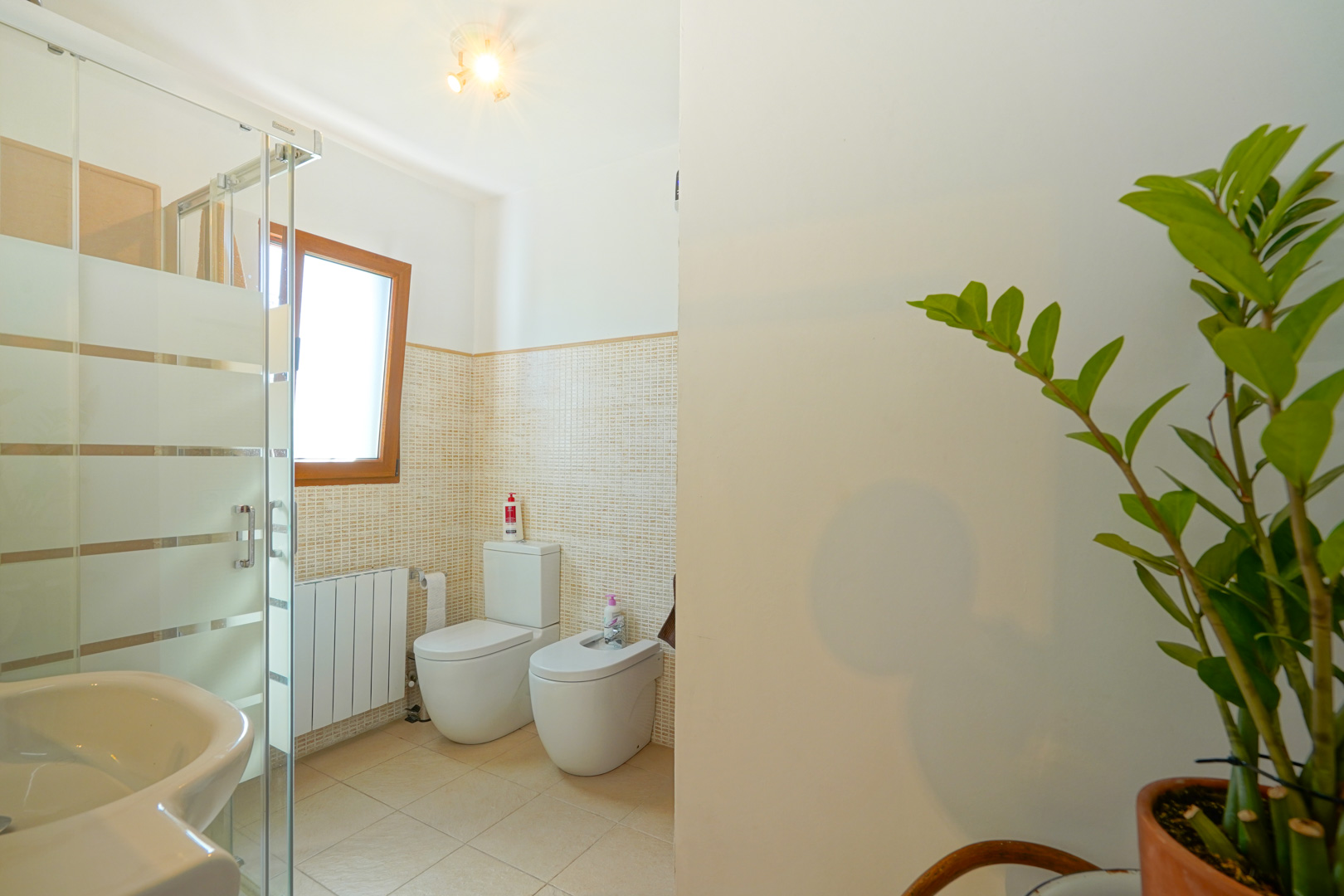 Bathroom featuring a glass-enclosed shower, toilet, bidet, and radiator, finished with beige mosaic wall tiles and a natural wood window frame.