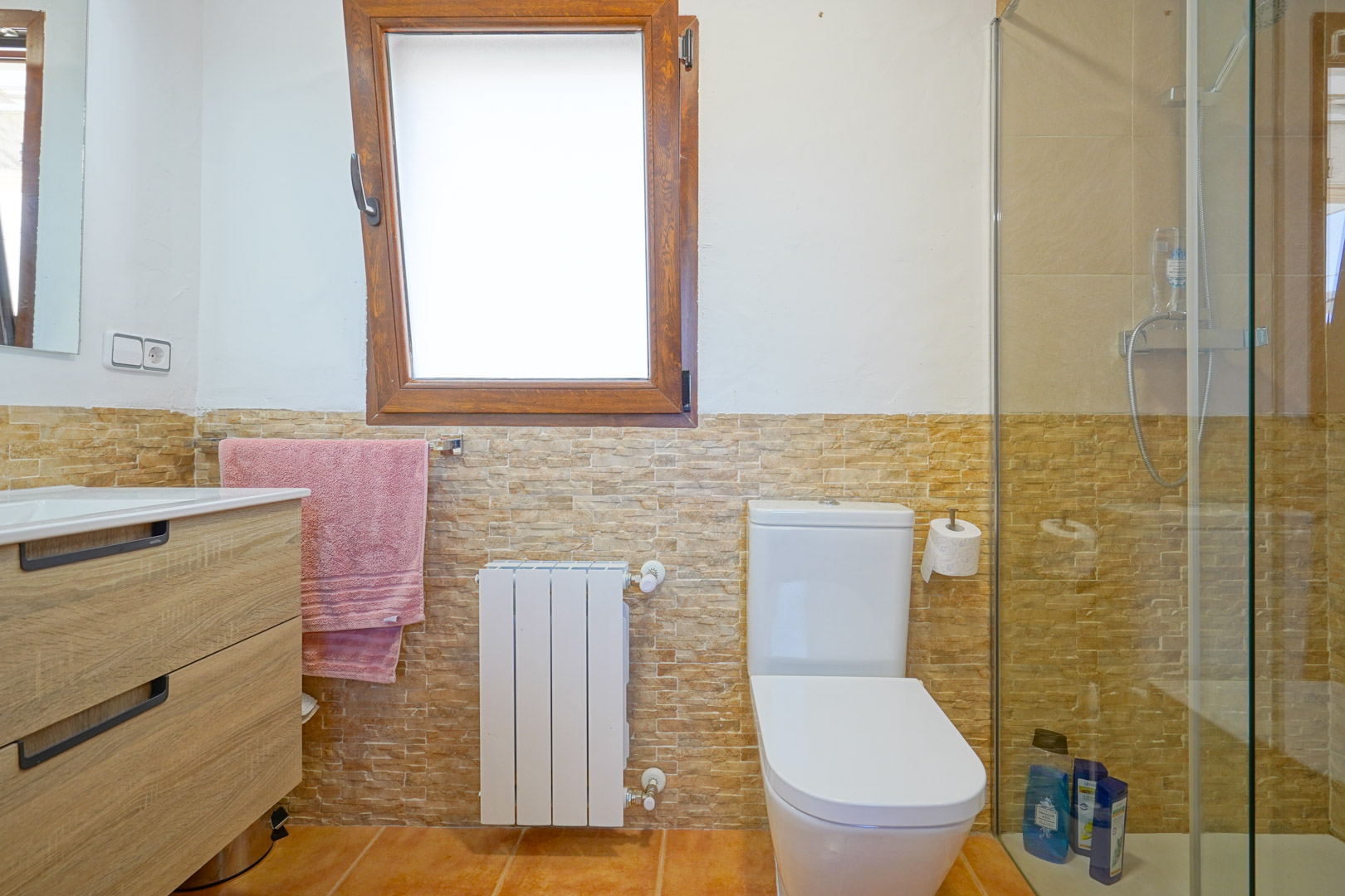 Bathroom featuring a wood-finish vanity, stone-effect wall tiling, a white radiator, and a walk-in shower with a glass partition next to a wooden window frame.