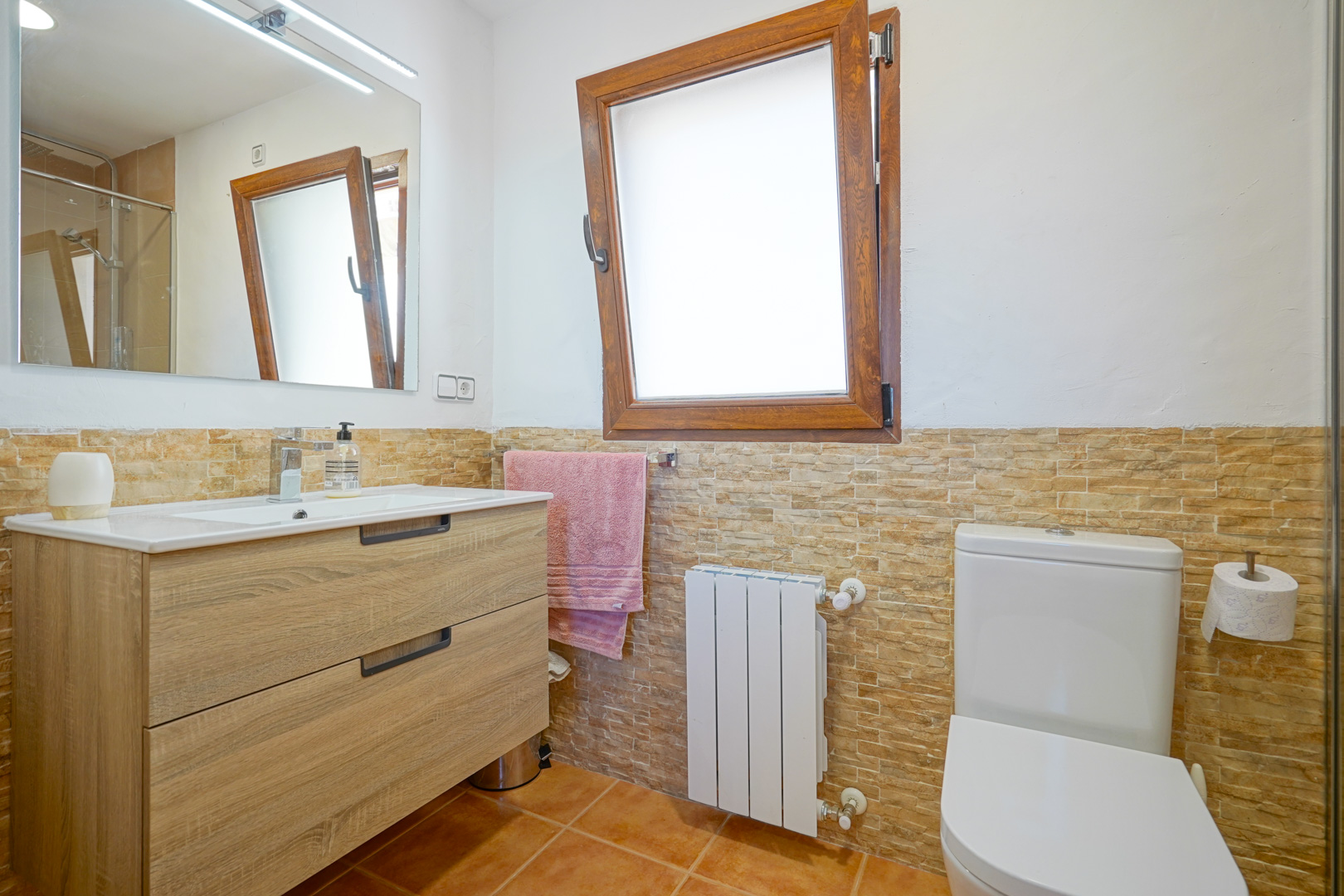 Bathroom featuring a wooden vanity with integrated sink, LED-lit mirror, and stone-effect wall tiling. Equipped with a radiator and a wooden tilt-and-turn window.