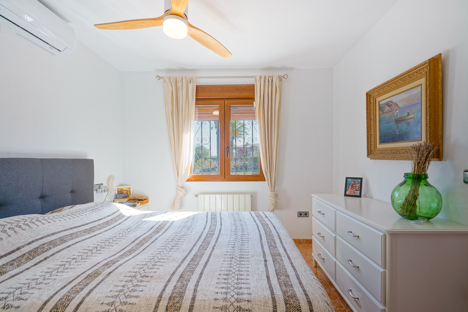 Bright bedroom featuring a wooden window frame, ceiling fan with wood blades, split air conditioning, and a white dresser. Includes radiator heating and tiled flooring.