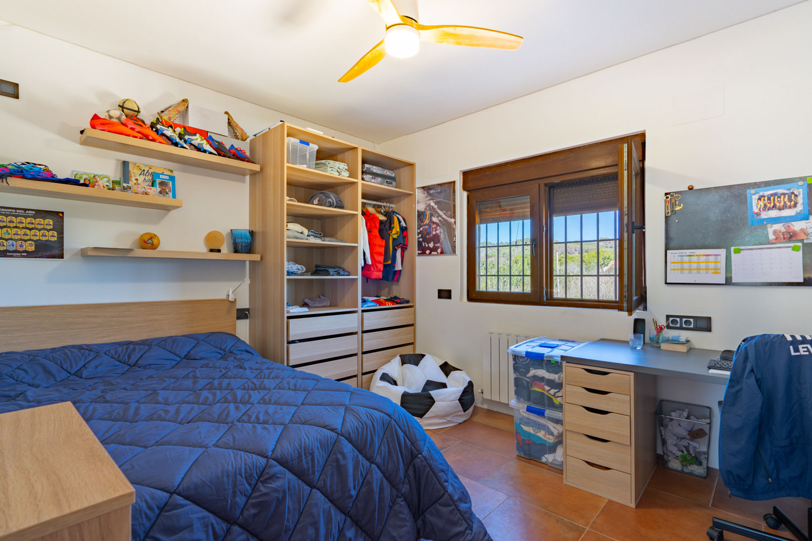 Bright bedroom featuring terracotta tile flooring, a built-in wooden desk, and open shelving. A large window with wooden frames offers views of the surrounding greenery.