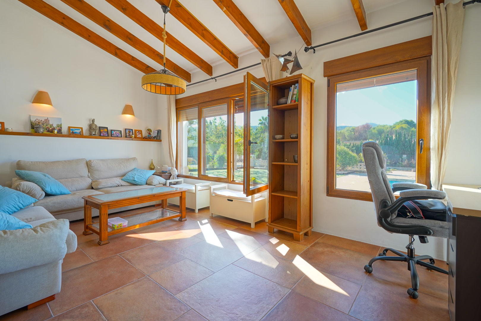Bright living area featuring exposed wooden ceiling beams, terracotta flooring, and a workspace. Large wooden-framed windows overlook the garden and pool.