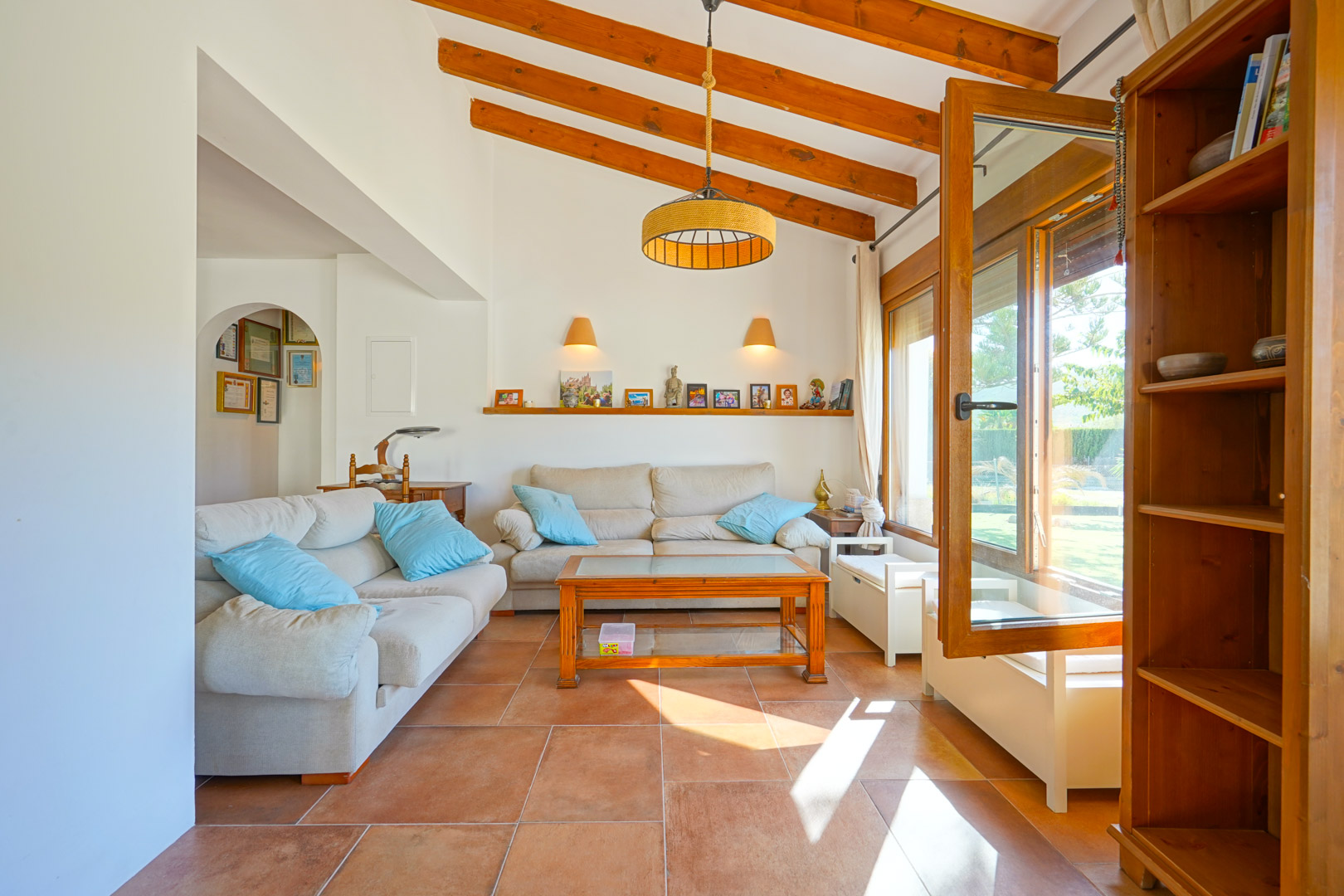 Bright living area featuring high ceilings with exposed wooden beams, rustic tile flooring, and large windows that invite natural light and garden views.