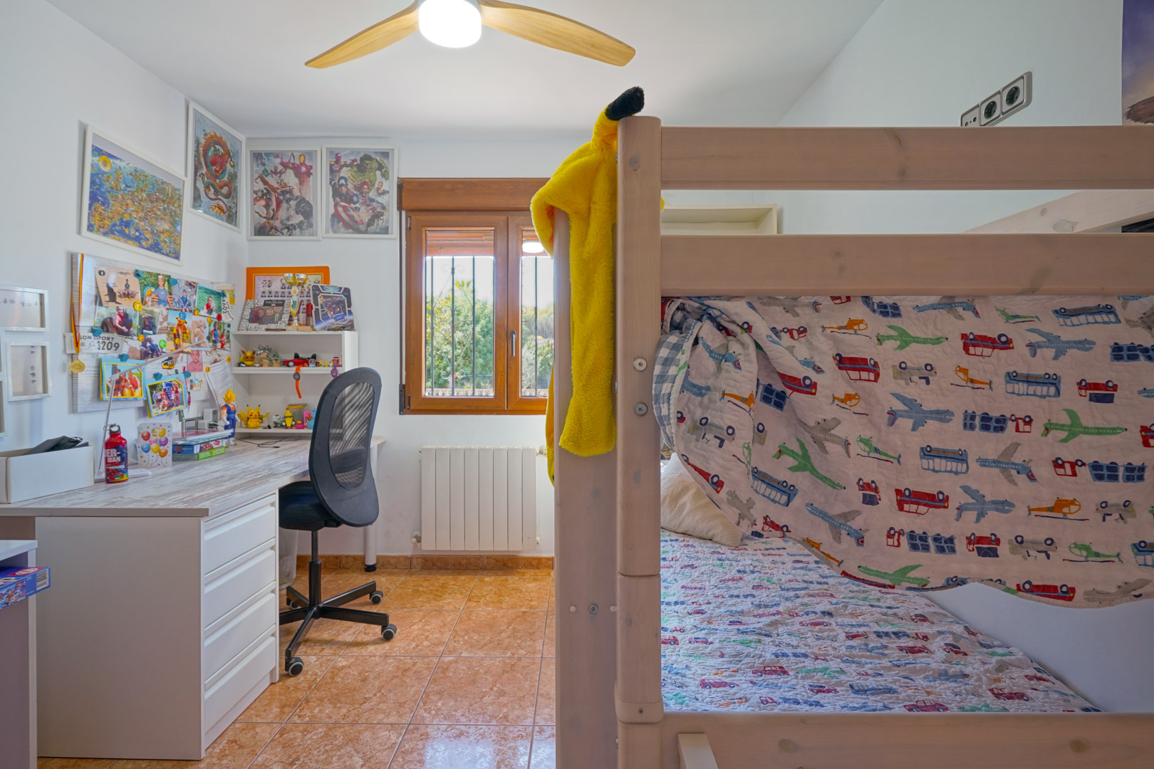 Children's bedroom featuring wooden bunk beds, a study desk, and a window with wooden frames. The room includes a radiator, ceiling fan, and tiled flooring.