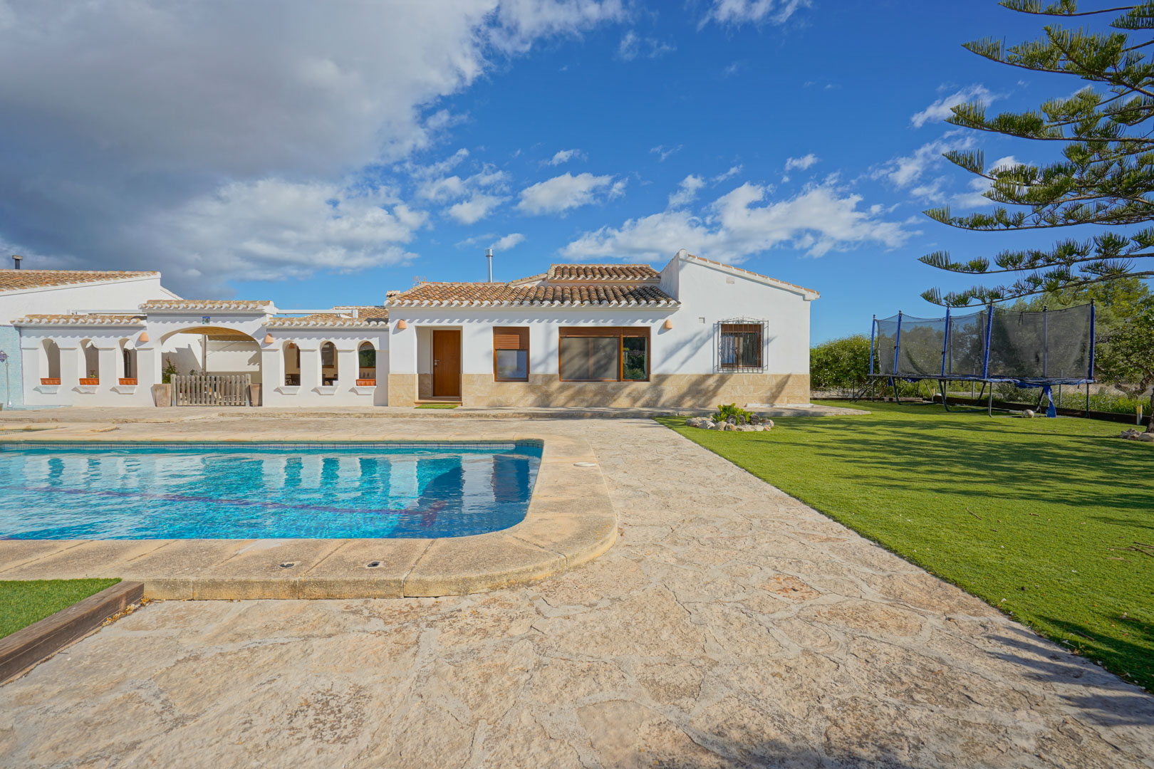 Exterior view featuring a large swimming pool, natural stone terrace, and manicured lawn adjacent to the white-walled villa with traditional terracotta roofing.