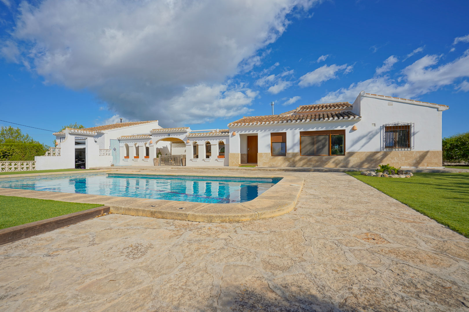 Exterior view of the villa featuring a private swimming pool, natural stone terrace, and manicured lawn areas under a bright sky.
