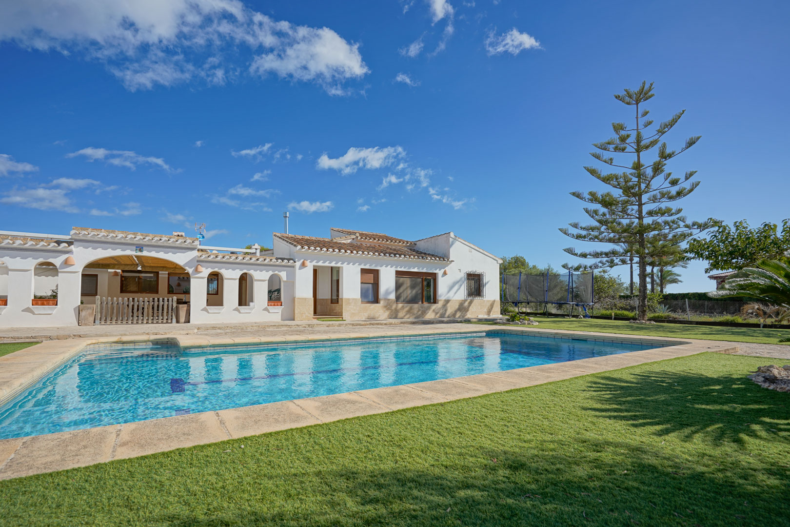 Exterior view of the villa featuring a rectangular swimming pool, manicured lawn, and stone terrace. The white-washed facade includes traditional arched porches and terracotta roofing.