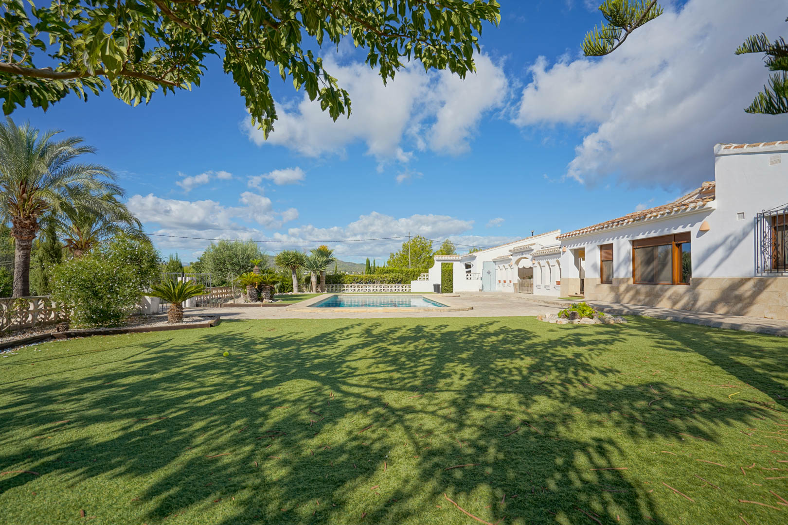 Exterior view of the white villa with stone accents, featuring a rectangular swimming pool, paved sun terrace, and a manicured lawn shaded by mature trees.