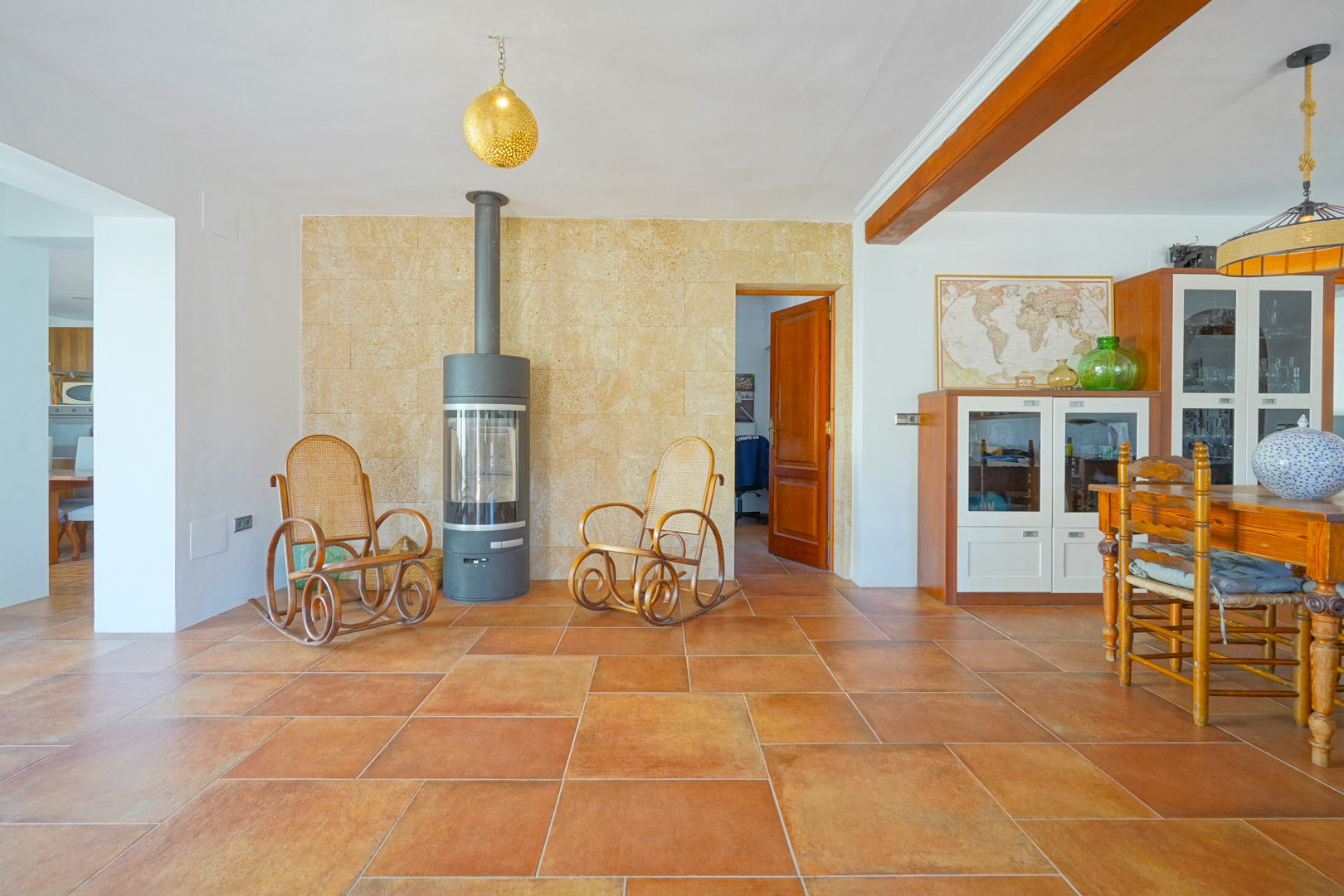Living-dining area featuring terracotta flooring, a modern wood-burning stove against a natural stone wall, and rustic wooden ceiling beams.