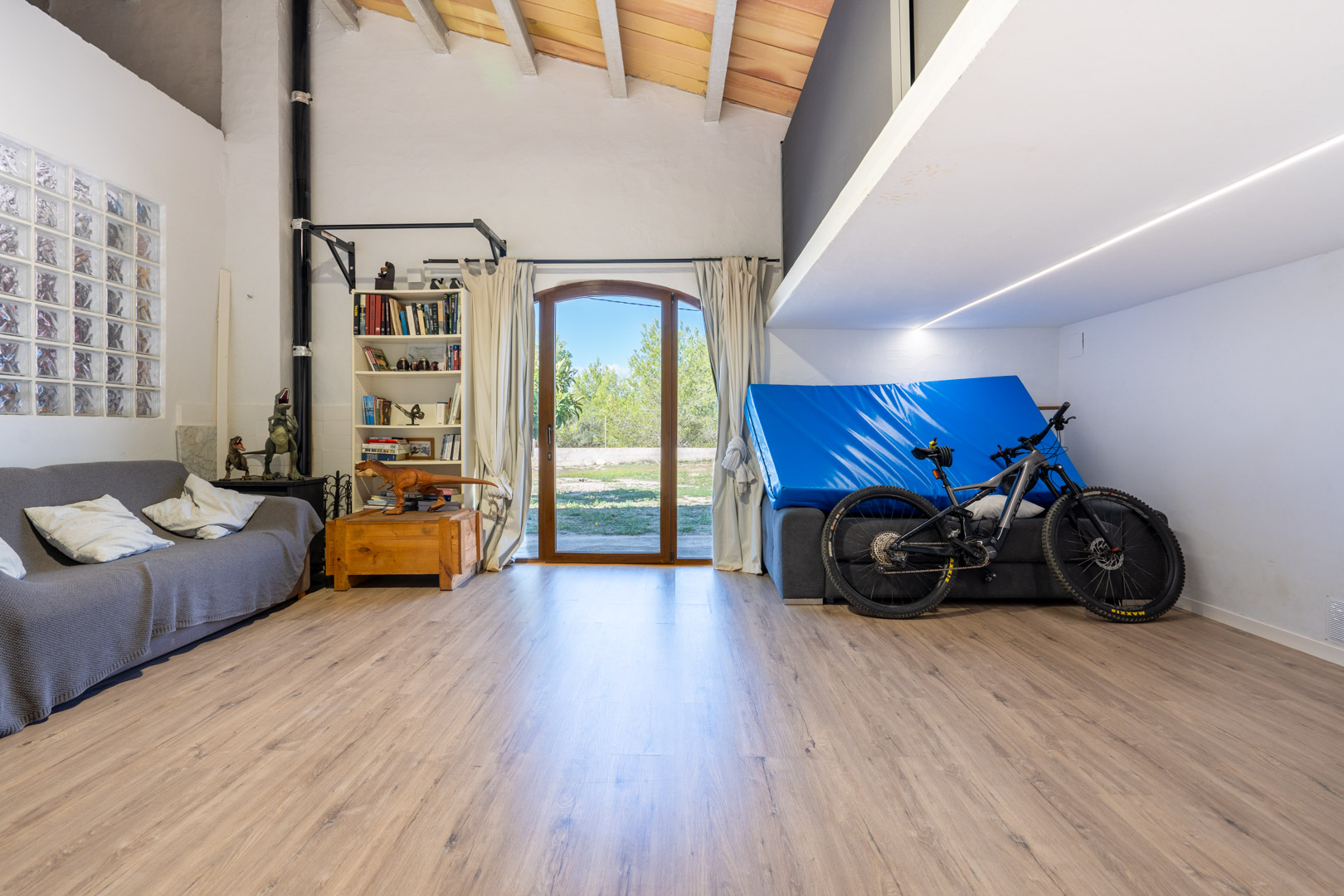 Multipurpose room featuring high vaulted ceilings with exposed beams, a mezzanine level, laminate flooring, and an arched glass door opening to the garden.