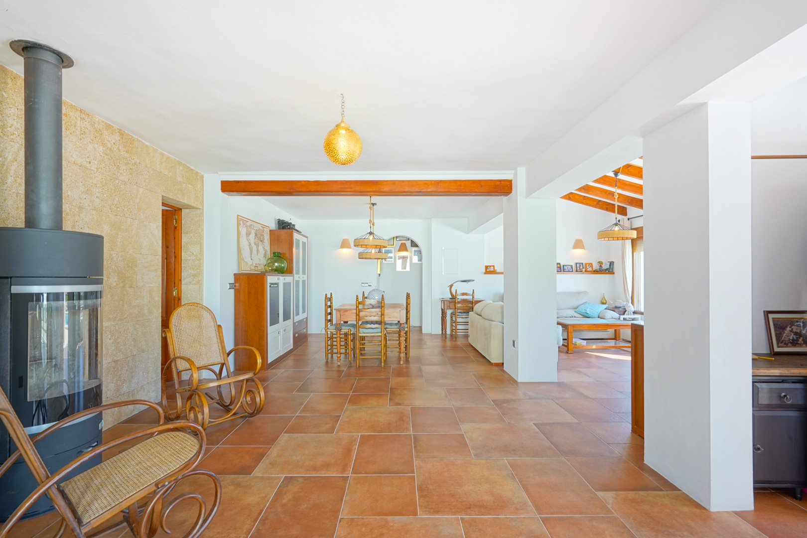 Open-plan living area featuring terracotta tile flooring, a modern wood-burning stove against a stone wall, and rustic wooden beams separating the dining space.