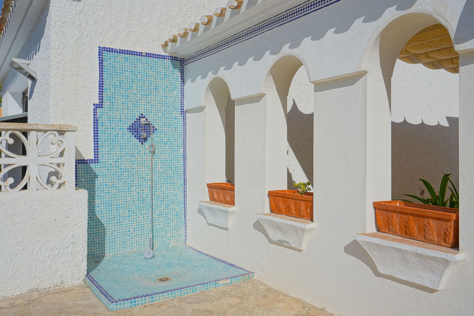 Outdoor shower featuring blue mosaic tiling set against a white facade with traditional Mediterranean arches and terracotta planters.