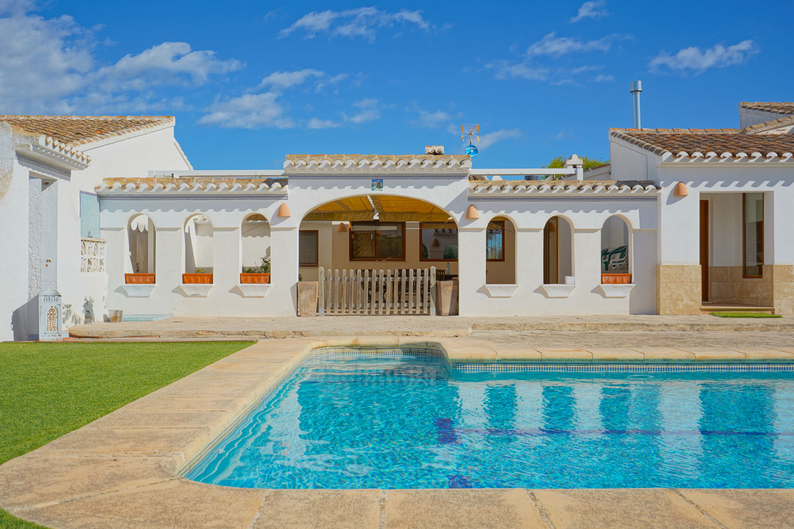 Outdoor swimming pool with natural stone coping and sun terrace facing a white Mediterranean facade featuring traditional arches and terracotta tile roofing.