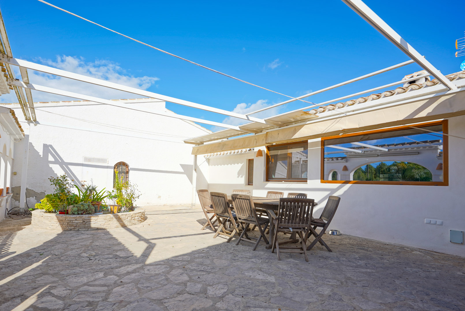 Outdoor terrace featuring irregular stone paving, a wooden dining set, and a metal pergola structure beside a circular stone planter.