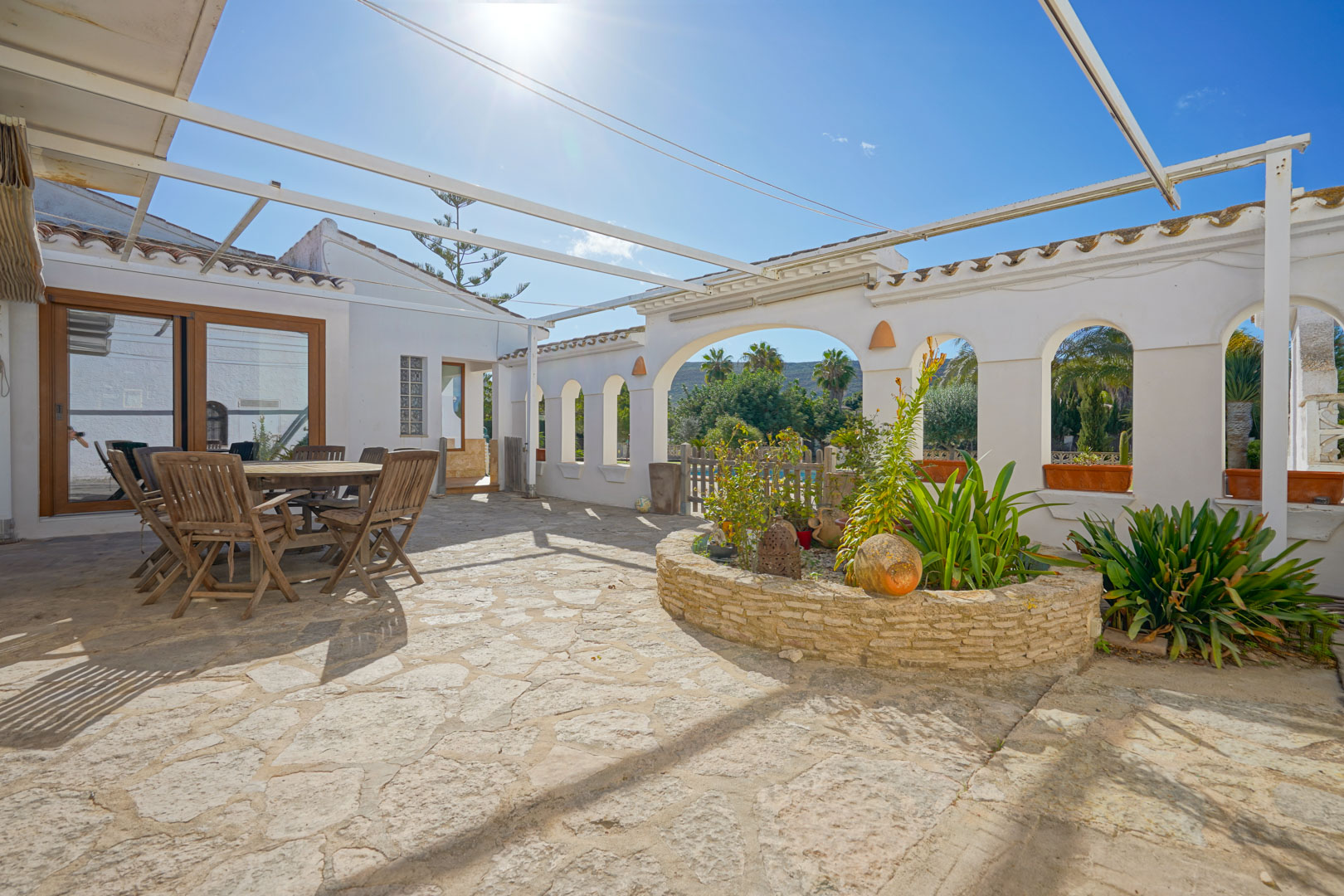 Outdoor terrace featuring natural stone paving, a wooden dining area, and a circular stone planter under a metal pergola structure.