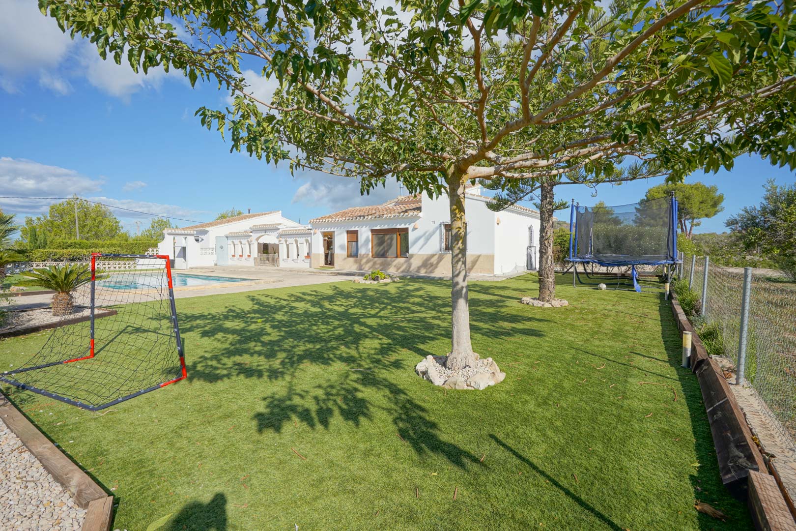Spacious garden with artificial turf and recreational area featuring a soccer goal and trampoline, set before the villa's white facade and swimming pool.