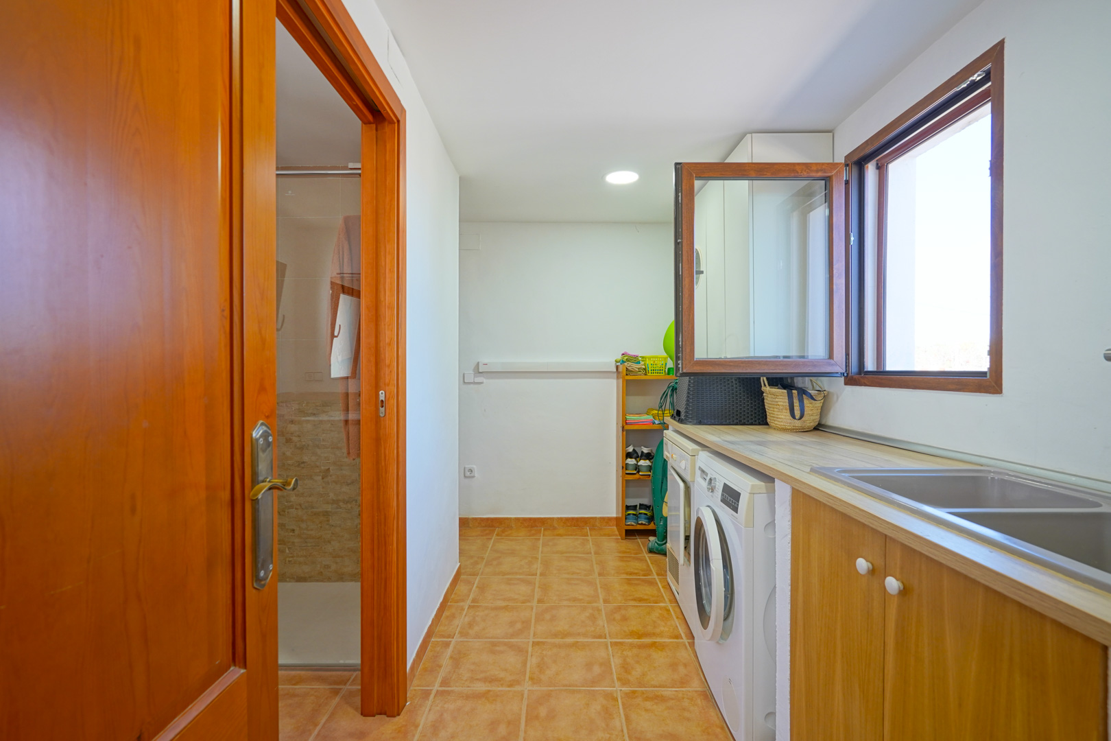 Spacious laundry room equipped with a washer, dryer, and double sink under a wooden countertop, with direct access to a bathroom.