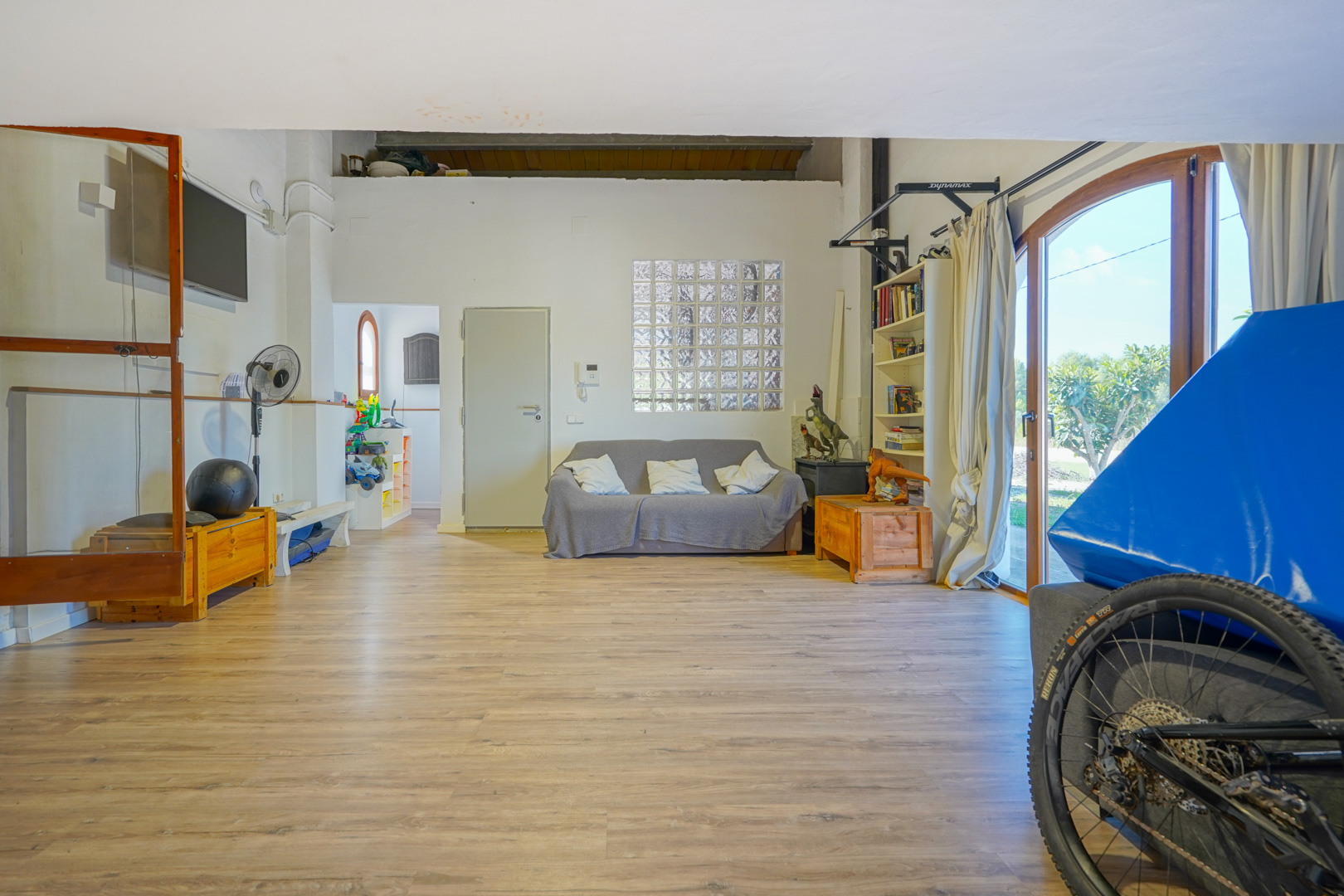 Spacious multipurpose room with wood-effect flooring, a lounge area, and direct outdoor access through large arched glass doors and glass block windows.