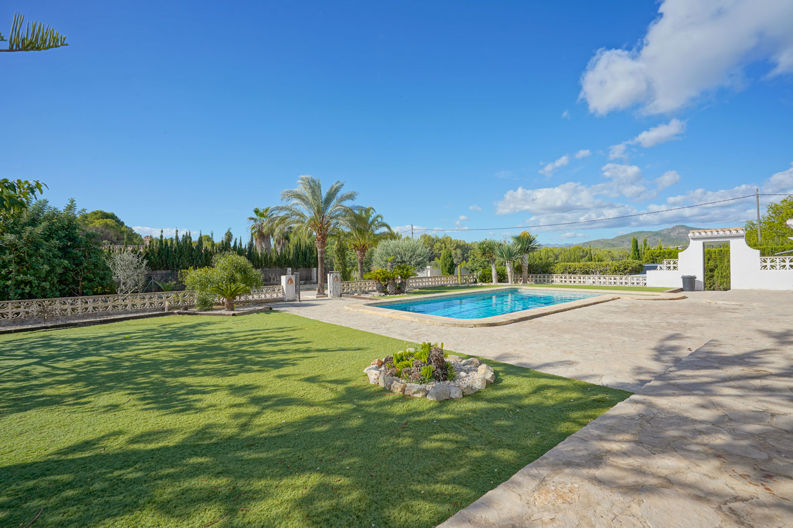 Spacious outdoor area featuring a rectangular swimming pool, natural stone sun terrace, and manicured lawn bordered by Mediterranean palm trees.