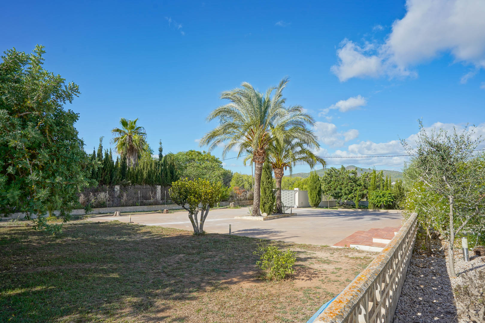 Spacious paved driveway featuring central palm trees, surrounded by Mediterranean greenery and views of the distant mountains under a clear sky.