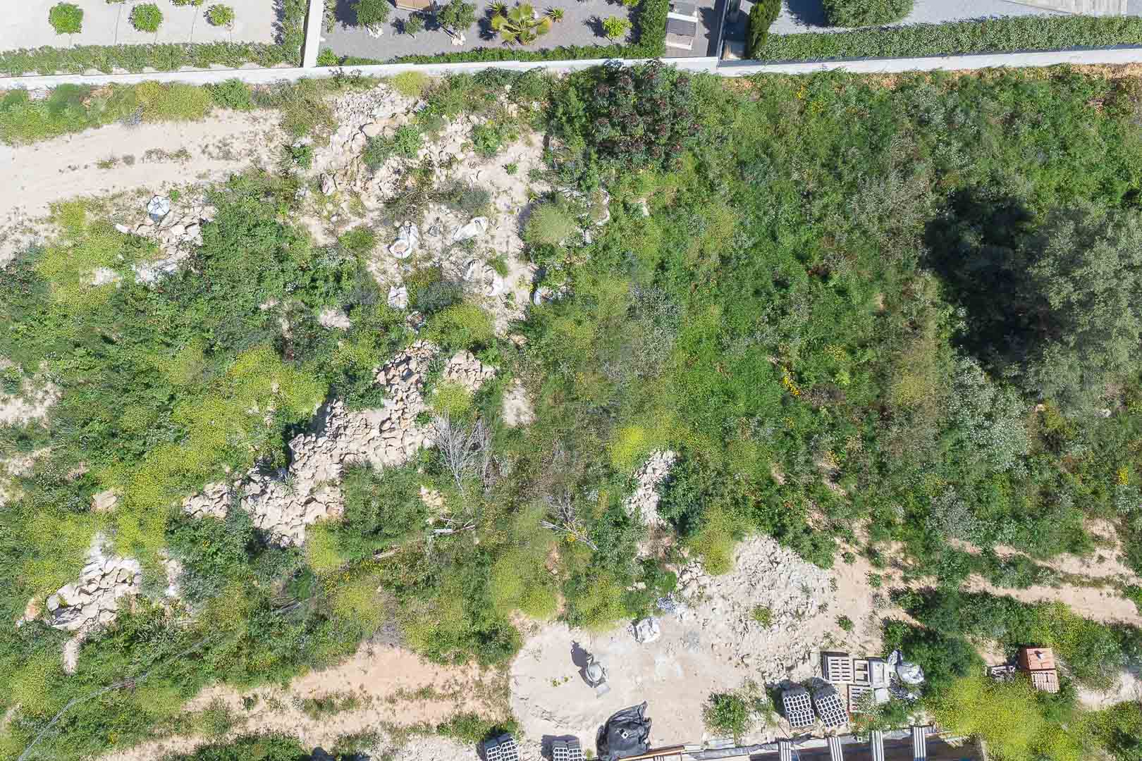 Overhead aerial view of the 1,000 m² plot, featuring natural Mediterranean terrain, rocky areas, and initial construction materials on-site.