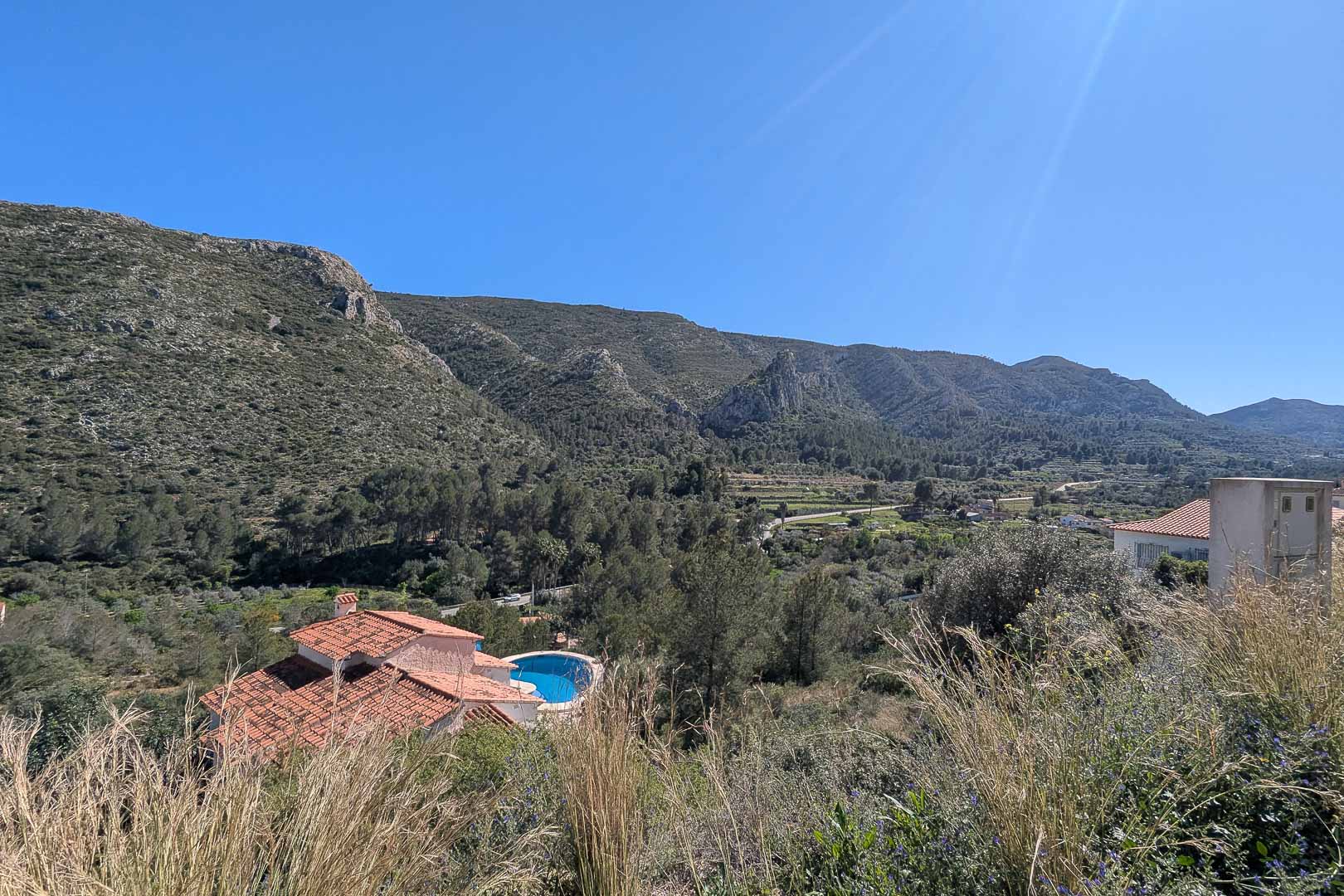 Elevated panoramic views overlooking the Pedreguer valley and rugged mountain range, featuring Mediterranean flora and a neighboring villa with a pool.
