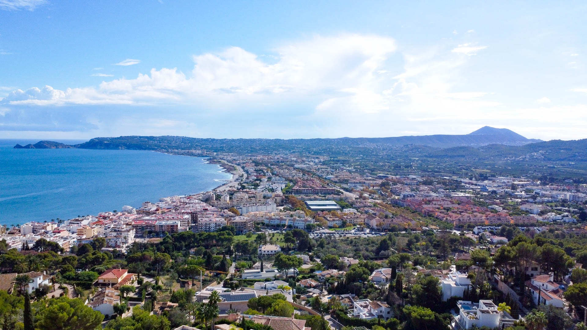Panoramic aerial view of Jávea Bay and the port from La Corona, featuring the Mediterranean coastline and the Montgó mountain in the distance.