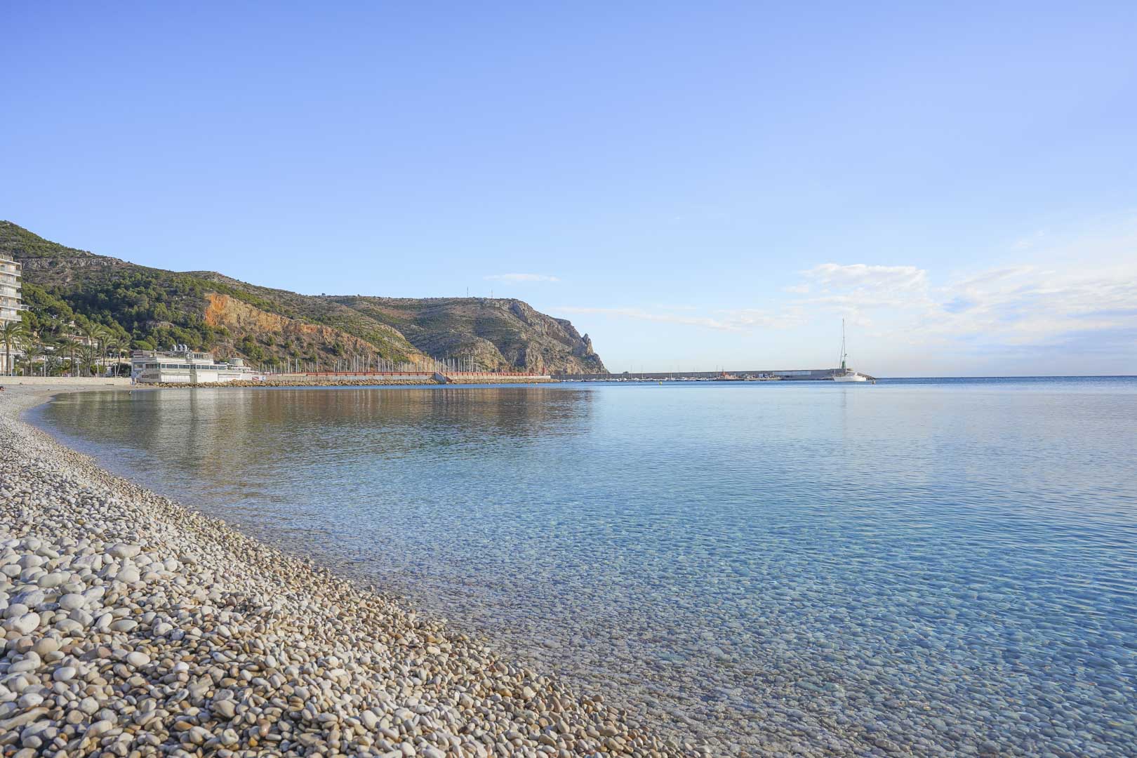 Panoramic view of the pebble beach and turquoise waters in Jávea, featuring the harbor breakwater and coastal cliffs under a clear blue sky.