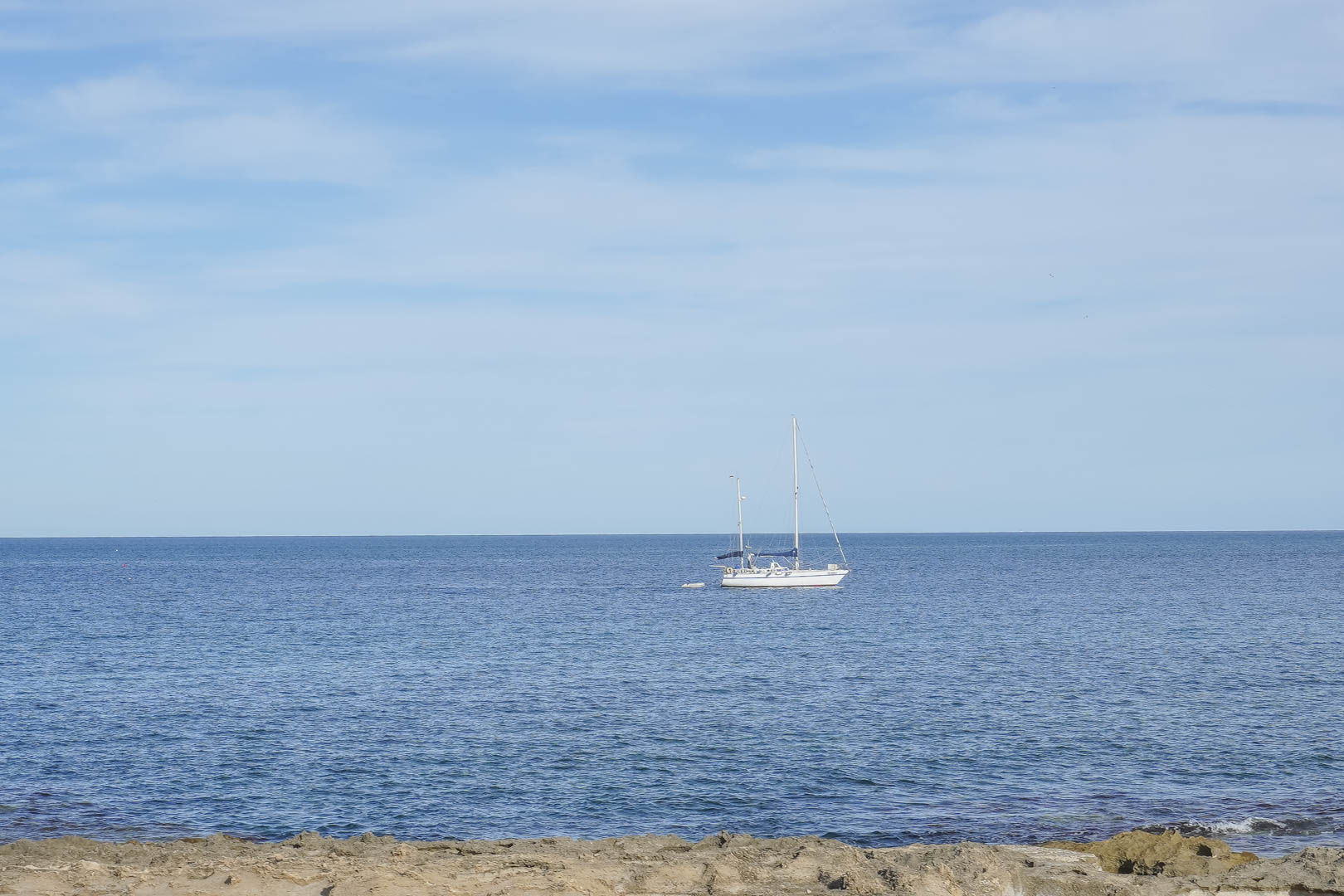 Panoramic views of the Mediterranean Sea from the Jávea coast, featuring a white sailboat anchored near the rocky shoreline under a clear sky.