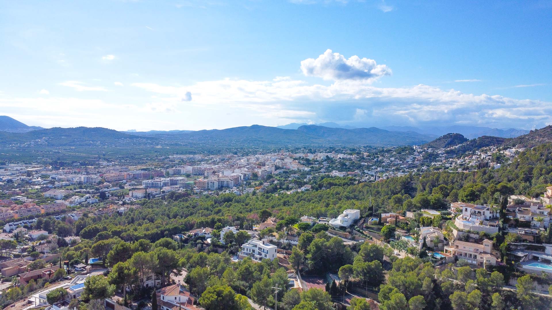 Aerial panoramic view of Jávea featuring the town center, dense pine forests, and the surrounding mountain range under a clear sky.