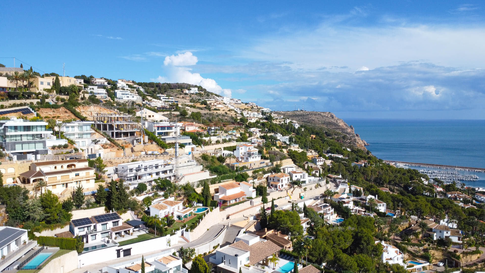 Aerial view of the La Corona hillside in Jávea, showcasing the terraced residential layout with expansive views of the Mediterranean coastline and the local marina.