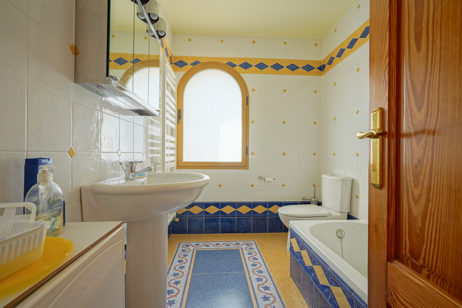 Bathroom featuring decorative blue and yellow tiling, an arched wooden window, and a functional layout with a pedestal sink, bathtub, and integrated towel radiator.